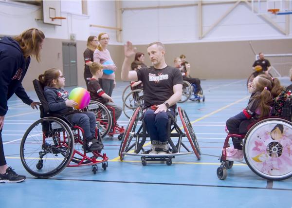 Rhyl Raptors COTM video story cover - 3 wheelchair users, 1 coach in the middle and 2 young girls each side, engaged in a wheelchair basketball training session on an indoor court.