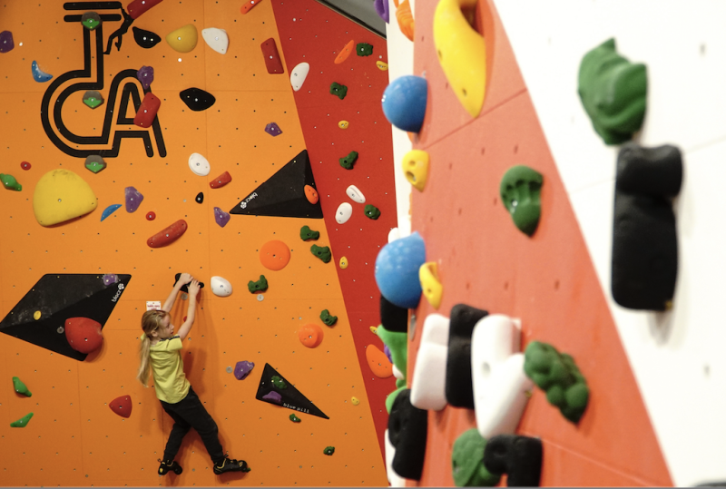 A small blonde child is bouldering on a distant wall. Climbing holds are in focus in the foreground on the right