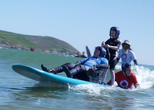 A young female adaptive surfer, sat on a seated board and catching a wave. Her younger sister is stood behind her on the board, smiling. Both are in wetsuits and water helmets, wearing buoyancy aids and blue UV vests over the top. They're smiling as the board catches the wave, with white water foaming around the back of the board, where a volunteer is lying off the back of the board and helping to steer the pair.