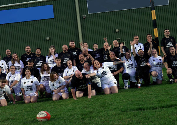 Dragons RFC Club of the Month September 2023 cover image - showing 3 of the Dragons adaptive teams stood and sat in rows together underneath the crossbars on a grass pitch. They are wearing a mixture of white or black strips, smiling, waving to the camera and laughing together.
