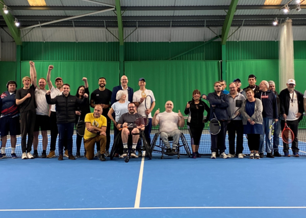 Club of the month cover shot, showing a range of participants stood and  sat in their wheelchairs in a line across an indoor tennis court, waving at the camera.