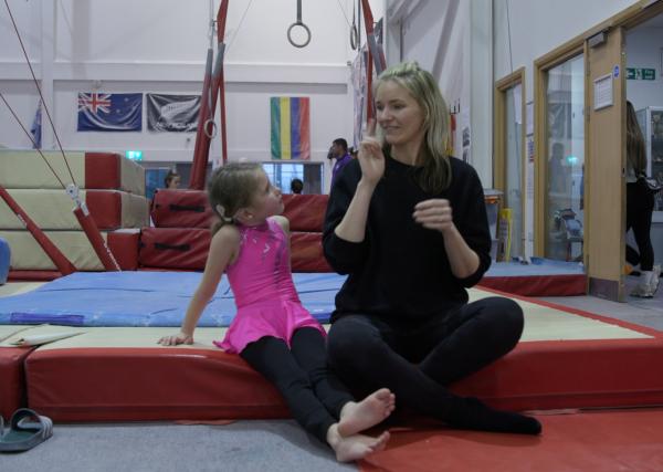 A mother and daughter, sat facing and signing to each other at an indoor gymnastics centre